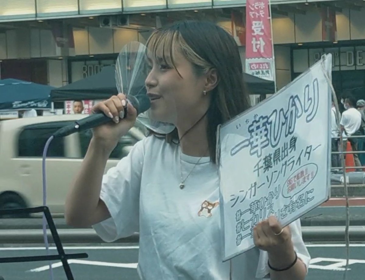 A street performer, identified by her sign as '一華ひかり' (Ichika Hikari) from Chiba Prefecture, singing into a microphone on a Japanese street.