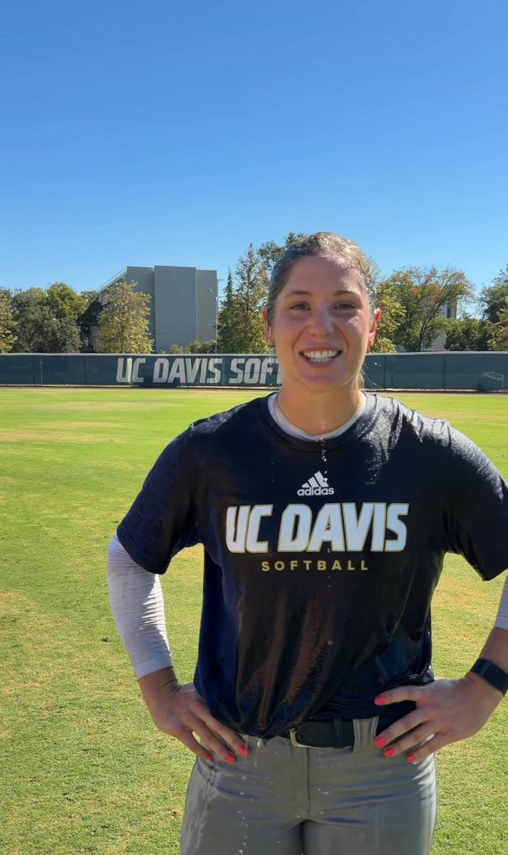A smiling woman stands on a grassy field wearing a black 'UC DAVIS SOFTBALL' Adidas shirt, with a UC Davis Softball banner visible in the background.