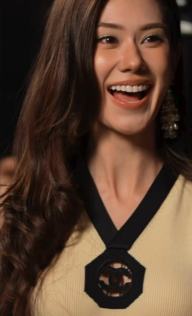 A smiling woman with long brown hair laughs joyfully, wearing a yellow and black sweater featuring a circular logo design.
