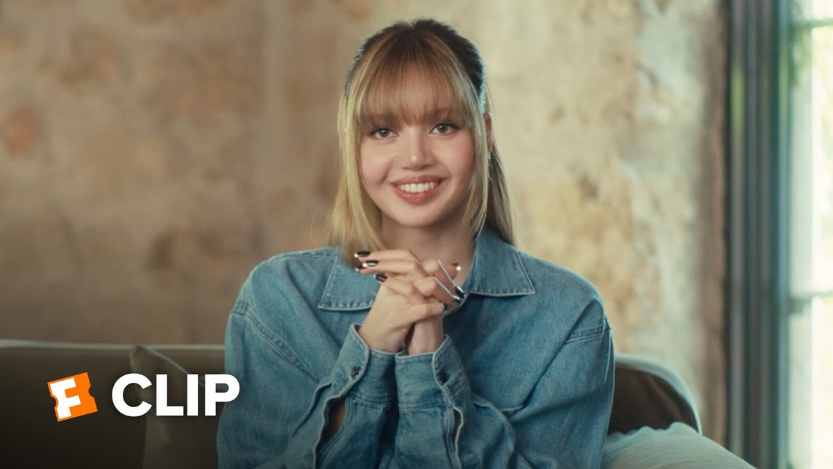 A smiling young woman with blonde bangs and a denim shirt sits on a couch in front of a stone wall, with a Fandom 'CLIP' logo visible in the corner.