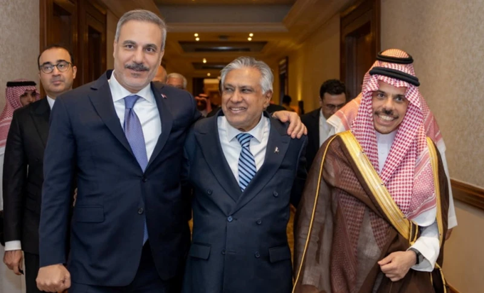Three men, including one in traditional Saudi attire and two in suits, stand together with their arms around each other, smiling in what appears to be a formal hallway setting.
