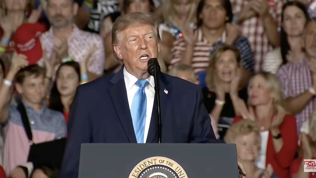 Donald Trump speaks at a Nebraska rally from behind a podium displaying the Presidential Seal, while a cheering crowd in red, white, and blue attire stands behind him; the post's reference to Natalie Harp joining him onstage relates to the rally context, though she is not visible in this particular frame.