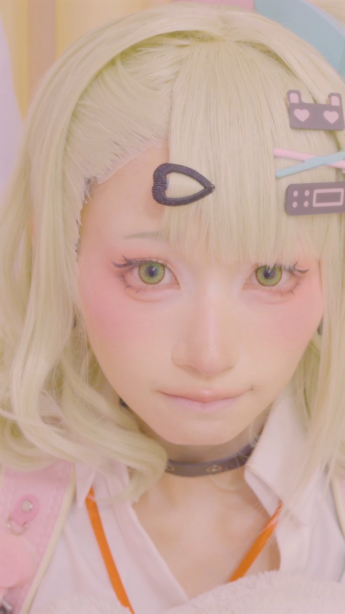 A cosplayer with blonde hair, green eyes, and pastel hair clips featuring heart and game console designs stares directly at the camera.