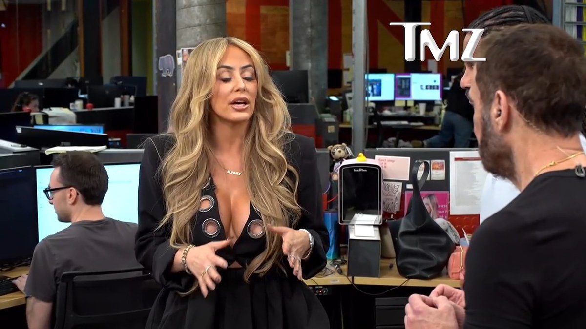 A blonde woman in a black cutout blazer gestures animatedly during an interview in the TMZ newsroom, telling a bearded man seated across from her to "Stay on his neck queen."