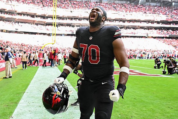 Football player number 70 of the Houston Texans, wearing a black uniform with a backwards cap, yells in celebration while holding his helmet on the field.