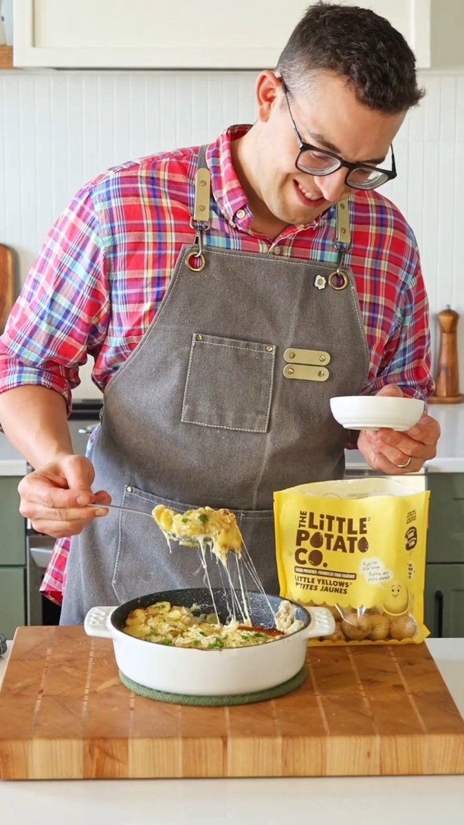 A man wearing glasses, a plaid shirt, and a gray apron smiling as he serves a cheesy potato dish from a white pan, with a bag of 'The Little Potato Co.' visible nearby.