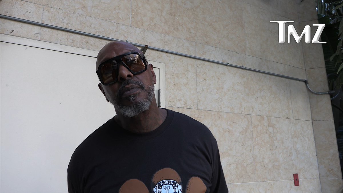 The image shows a bald Black man with a gray goatee wearing reflective sunglasses and a black graphic t-shirt featuring "The World" text, standing outside against a beige tiled wall. The TMZ logo is visible in the upper right corner, suggesting the photo accompanies a TMZ article about Sleepy Brown, the musician, expressing his strong opinion that critics should "shut the hell up" regarding Hip-Hop's inclusion in the Rock & Roll Hall of Fame.