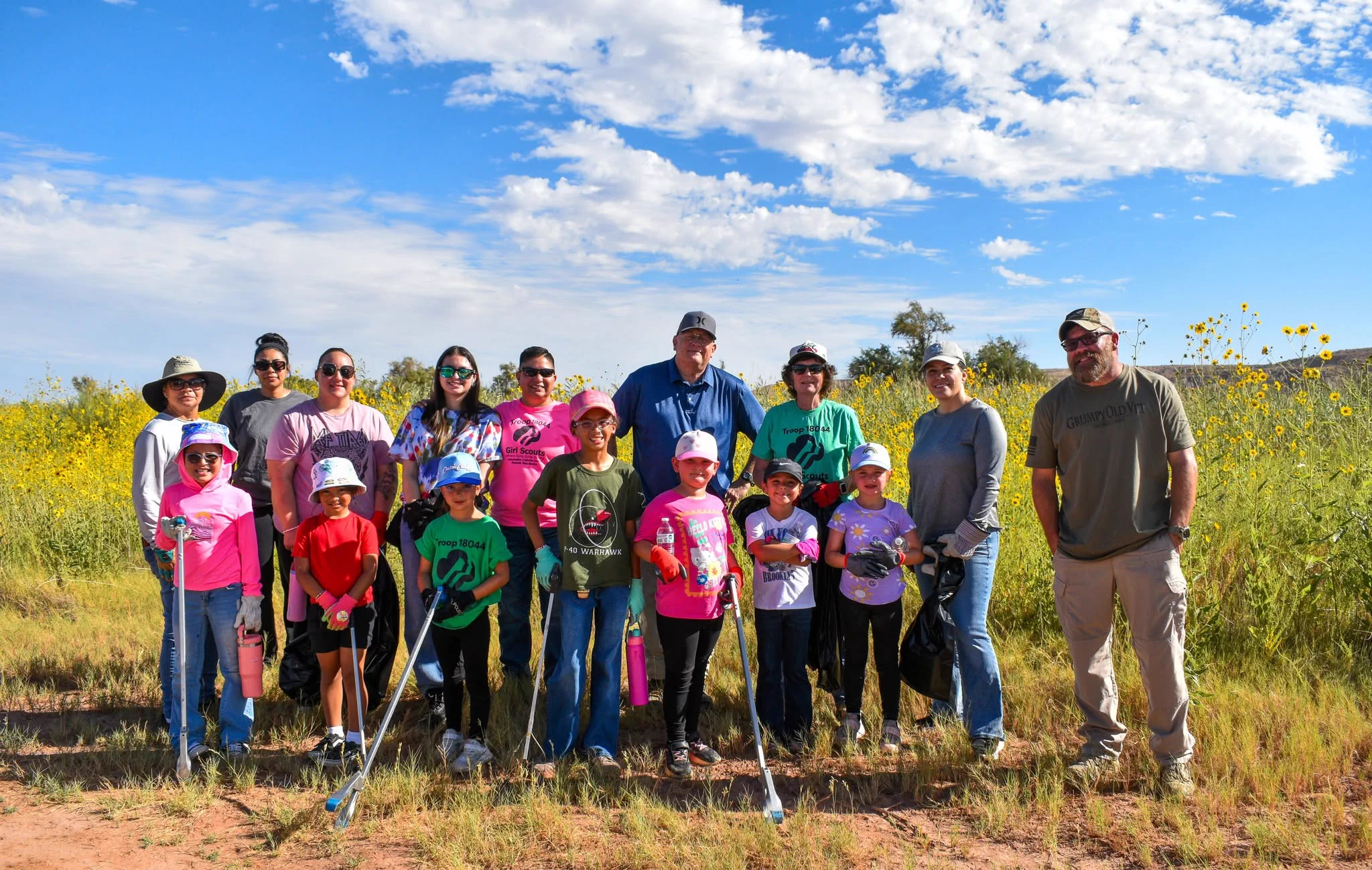 Group of adults and children standing outdoors in a sunny field holding trash pickers for a cleanup event.