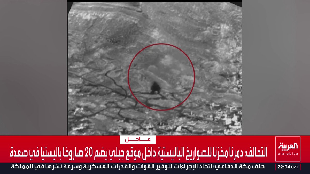 The image, broadcast on Al Arabiya news with a timestamp of 22:04 GMT, shows a grainy black-and-white aerial reconnaissance photo with a red circle highlighting a targeted site. Arabic text at the bottom reads "Urgent: Coalition destroys ballistic missile storage facility inside a Houthi site with 20 ballistic missiles in Saada," along with a related headline regarding Houthi military procedures. The photo appears to depict the aftermath or targeting of the missile storage facility referenced in the breaking news report.