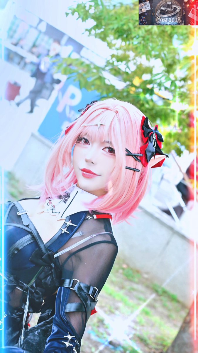 A cosplayer wearing a pink wig and a black gothic outfit poses outdoors at what appears to be a convention, with a 'COSFOCUS' watermark in the top right corner.