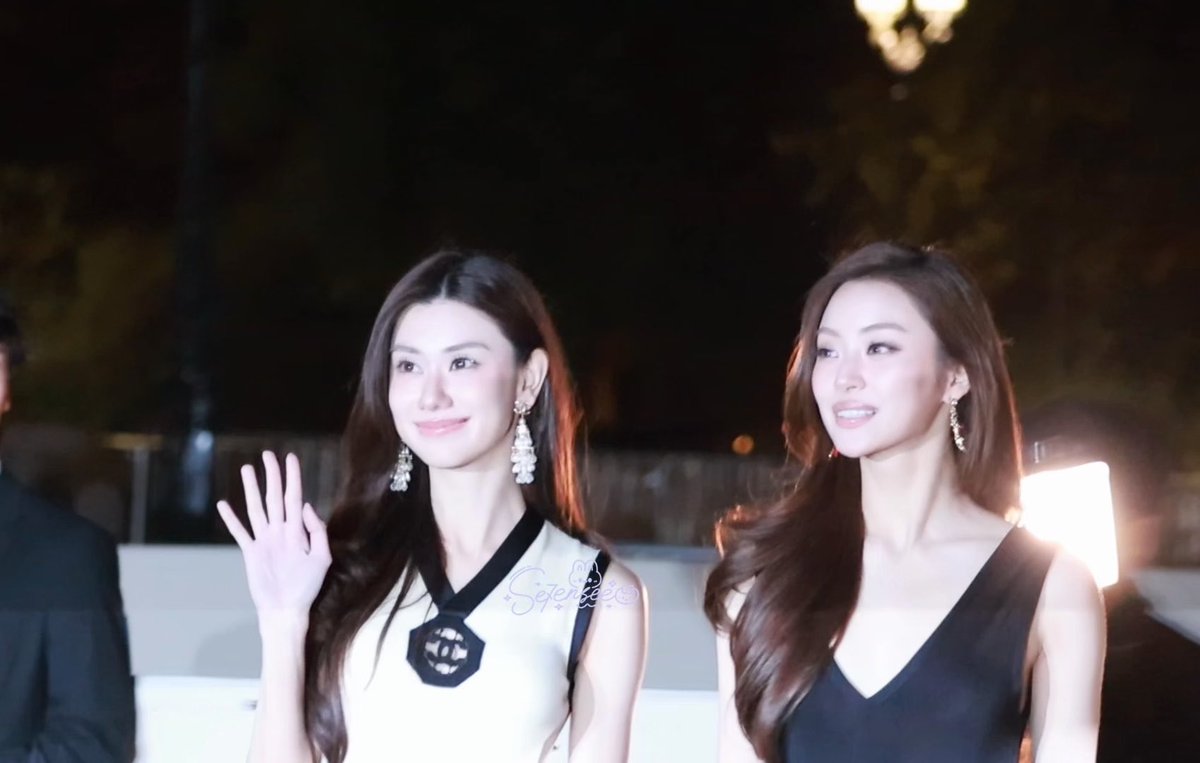 Two women at an evening event, one waving while wearing a white and black top with crystal earrings, the other in a black v-neck dress.