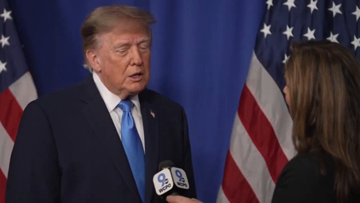 A man in a dark suit and blue tie is being interviewed by a reporter from WCPO Channel 9, with American flags displayed behind him. The image captures Trump during the interview referenced in the post, where he discussed a housing development in Ohio.