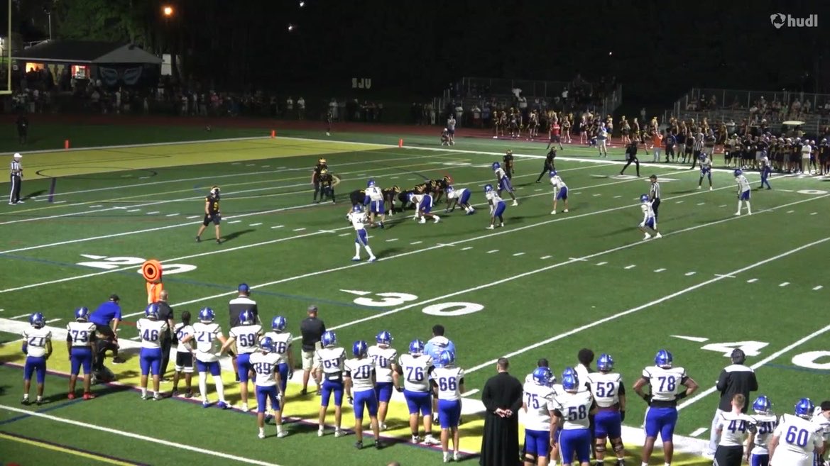 A nighttime high school football game scene on a turf field, with players in white and blue uniforms on the sidelines and a play in progress; 'hudl' watermark visible in the upper right corner.