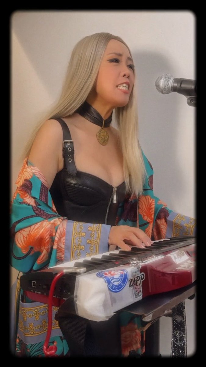 A blonde woman wearing a black leather corset top, choker, and teal patterned shawl sings into a microphone while playing a red keyboard adorned with stickers including 'ZAPP'.