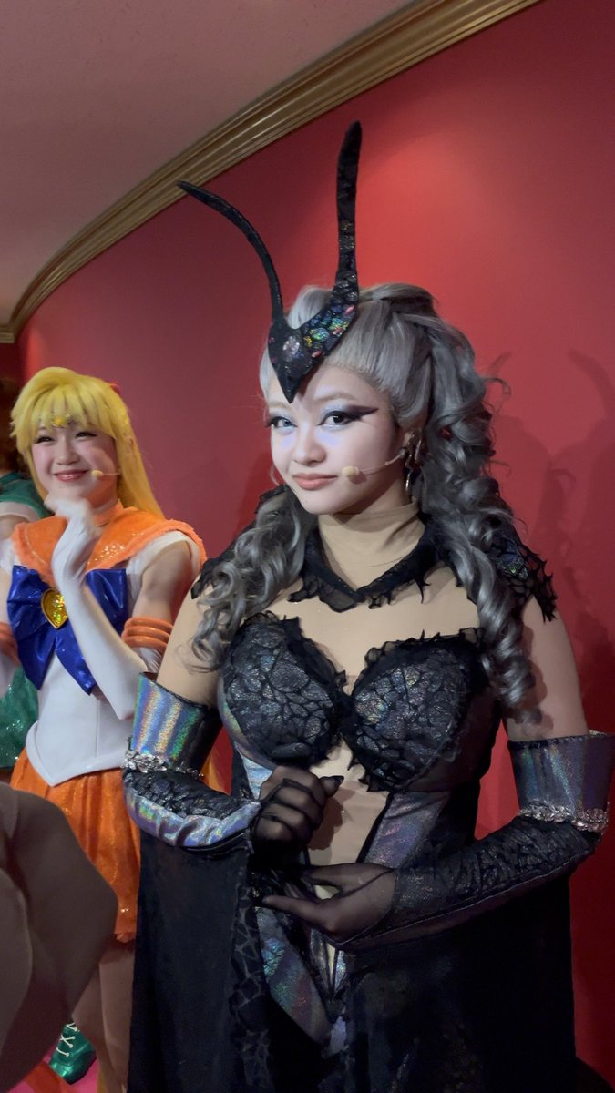 A young woman in an elaborate black theatrical costume with horn-like headgear, lace details, and shimmering sleeves, standing on a stage next to another performer dressed as Sailor Venus.