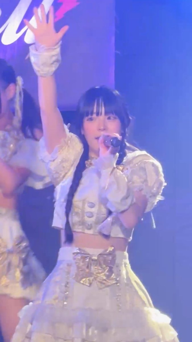 A young female performer in an ornate cream-colored ruffled idol costume sings into a microphone while raising her hand on a blue-lit stage.