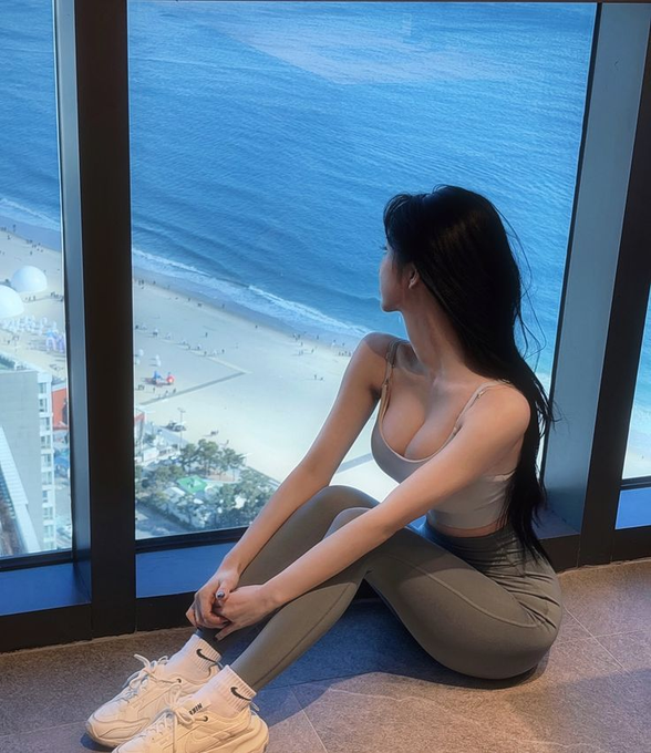 A woman with long black hair wearing a white crop top and grey leggings sits by a large window looking out at a beach and ocean.