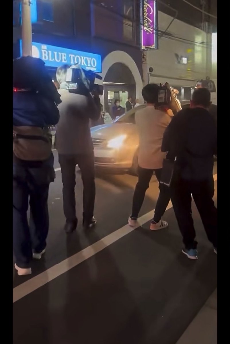 A nighttime street scene showing several people with cameras and equipment standing on a road in front of a venue with a blue 'BLUE TOKYO' sign, with a car passing by.