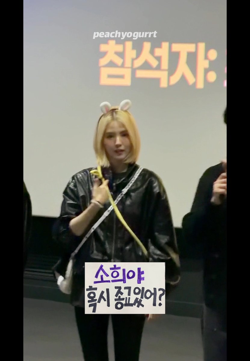 A screenshot of a video showing a blonde woman with bunny ears wearing a black jacket, with Korean text '참석자:' behind her and a caption box reading '소희야 혹시 중근왔어?' attributed to 'peach yogurrt'.