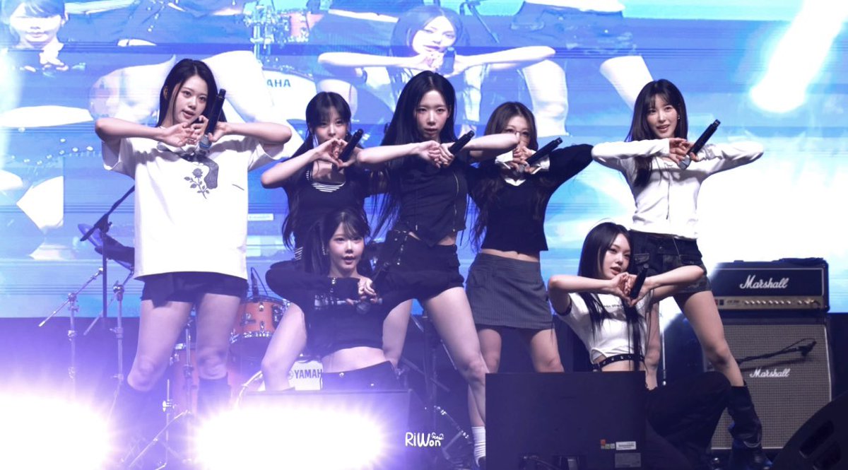 A K-pop girl group performing on stage with synchronized arm poses, holding microphones, with a Marshall amplifier visible on the right and a Yamaha drum kit in the background; the image is watermarked 'RIWO'.