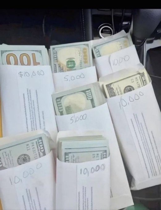 Several envelopes containing stacks of US hundred-dollar currency are labeled with handwritten amounts like '$10,000' and '5,000' on what appears to be a car seat.