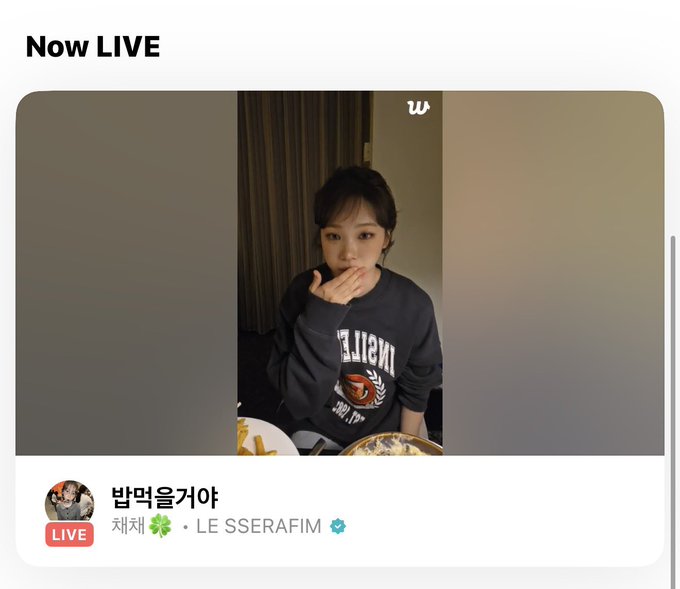 The image shows a screenshot of a Weverse LIVE broadcast featuring LE SSERAFIM member Kim Chaewon, with her username "밤먹을거야" (roughly meaning "I'll eat tonight") displayed alongside her stage name and group. She is seen wearing a black sweatshirt with a logo design, sitting at what appears to be a table with food in front of her, while her hand covers part of her face. The "Now LIVE" header and verified Weverse account indicators confirm this is the live streaming moment referenced in the post about Chaewon's broadcast.