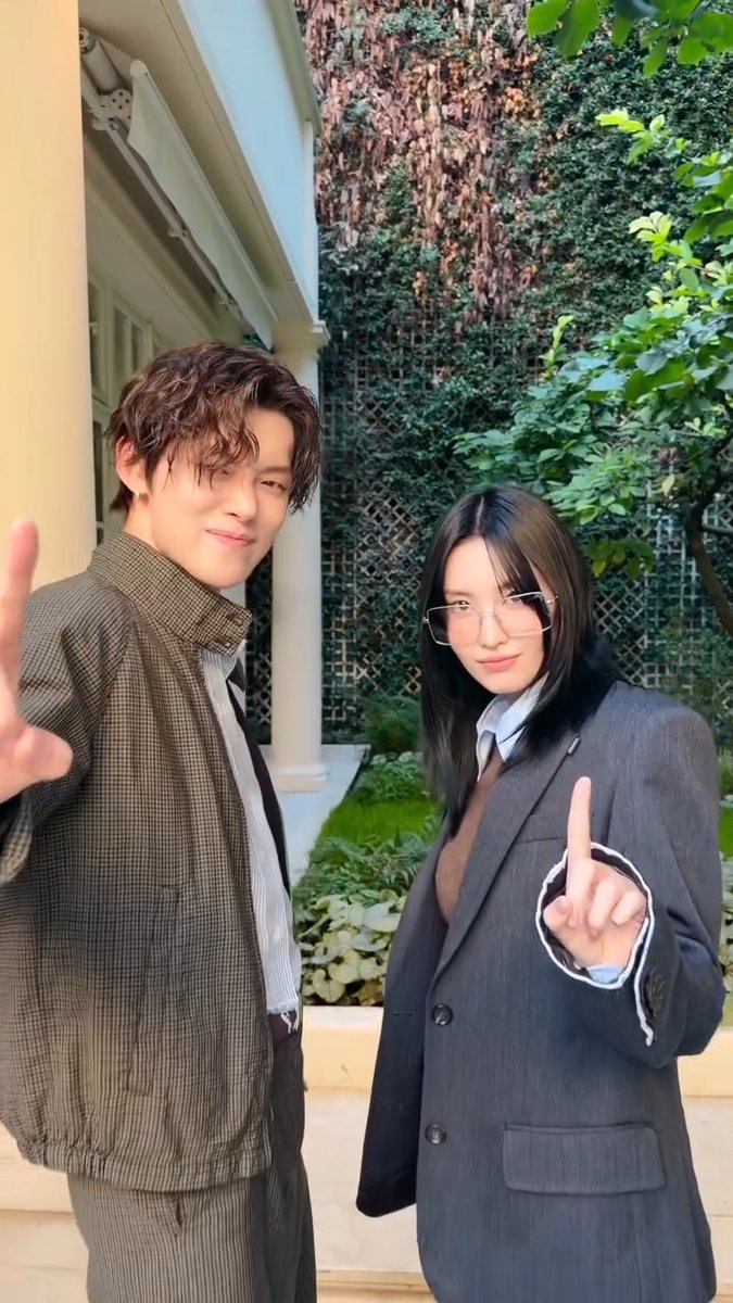 Two young people wearing suits pose outside; the male on the left with curly hair in a houndstooth jacket gives a peace sign, while the female on the right wears glasses and a dark blazer pointing upward.