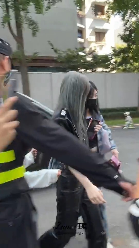 A blurry candid shot of people walking outdoors, including a woman with silver hair and another wearing a black mask, while someone in a security uniform reaches across the frame.