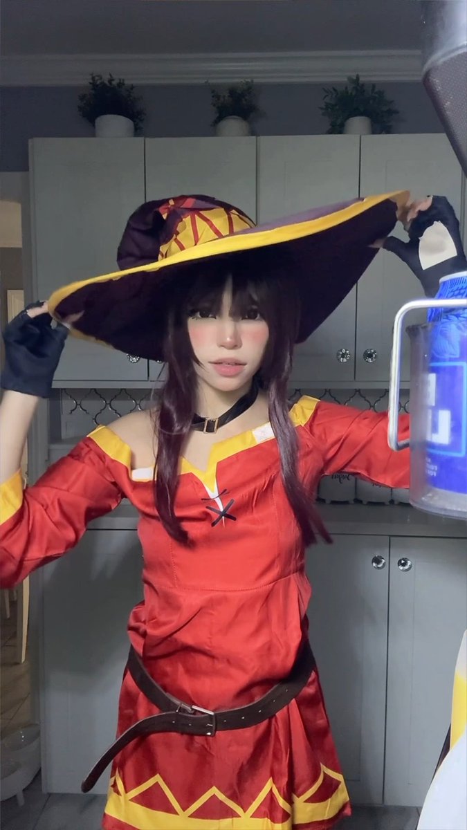 A person with dark hair poses in a red and yellow costume with a large witch-like hat and a black belt, holding a blue water jug in what appears to be a kitchen.