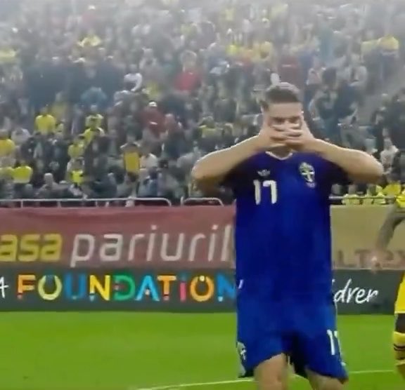 A soccer player wearing a blue Sweden kit (#17) stands on the field and makes a heart shape with his hands in front of his chest, while a crowded stadium is visible in the background behind advertising signs that read 'asa pariuri' and 'FOUNDATION'.