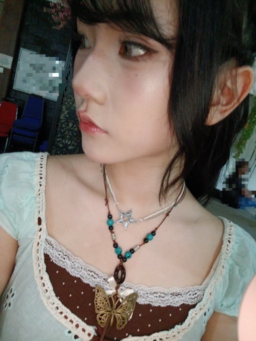 A close-up selfie shows a young woman with short bangs wearing a brown and cream top with lace trim, accessorized with a butterfly pendant necklace.