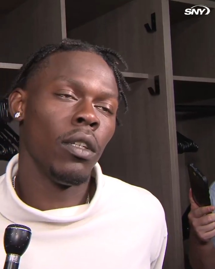 A close-up of a young Black man with braided hair being interviewed in a locker room, wearing a white shirt and an earring, with an 'SNY' logo in the corner.