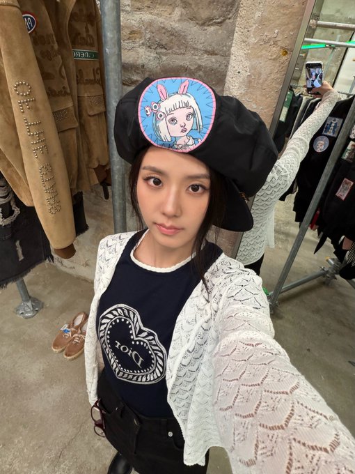 A selfie of a young woman wearing a black beret with a cartoon bunny patch, a navy 'Dickies' tank top, and a white lace cardigan in what appears to be a vintage clothing store.