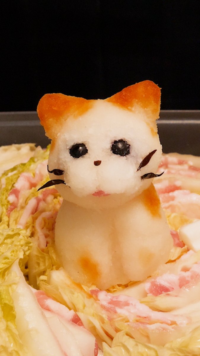 A close-up of a hot pot dish featuring a cute cat-shaped food item made from white and orange ingredients, sitting in a cabbage and meat broth.
