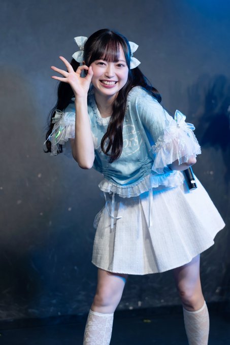 A smiling young woman with long dark hair in pigtails, wearing a blue and white ruffled outfit with hair bows, posing playfully against a dark background.