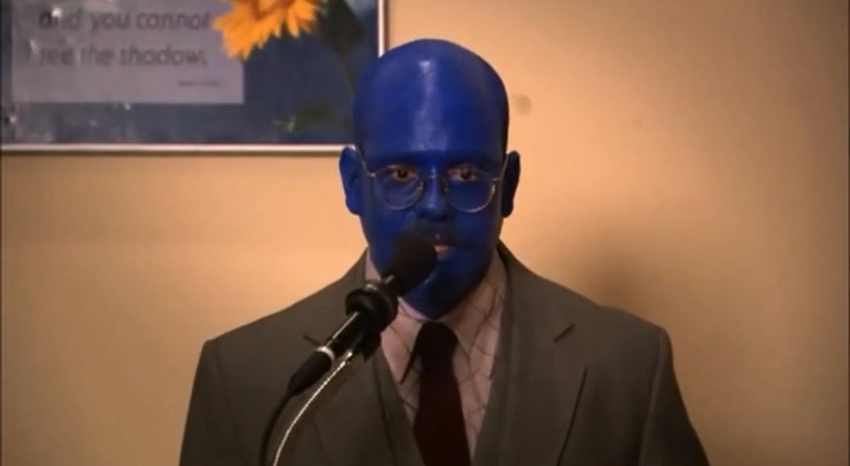 A man with a blue-painted head, glasses, and a gray suit speaks into a microphone in front of a sunflower poster that reads "and you cannot see the shadows," capturing the hopeful optimism of a long-suffering Cleveland Guardians fan heading into the postseason.
