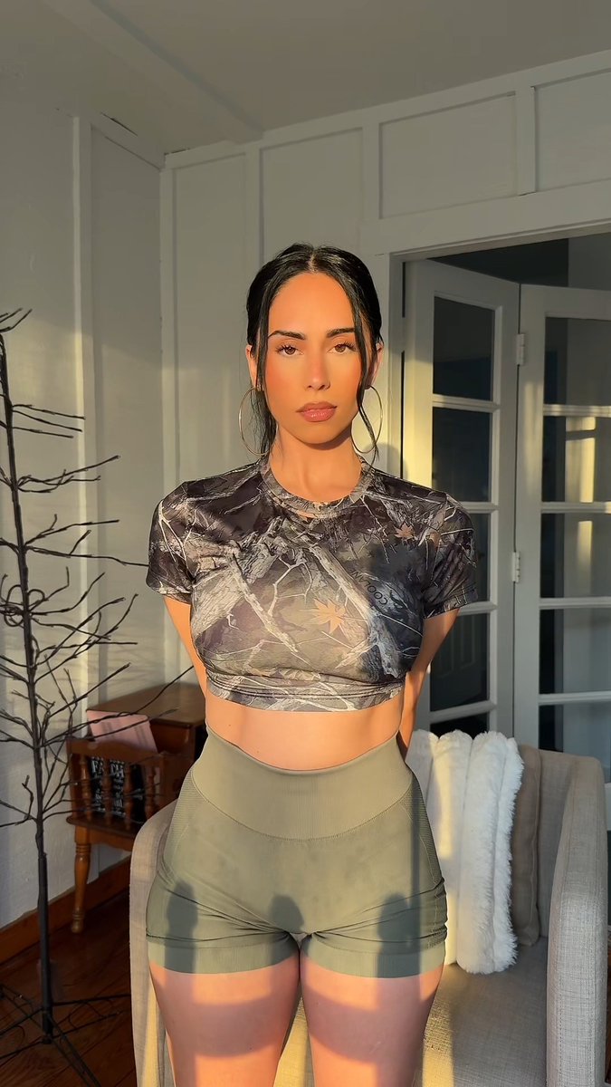 A woman stands indoors wearing a camouflage crop top and olive green athletic shorts, illuminated by sunlight near a window.