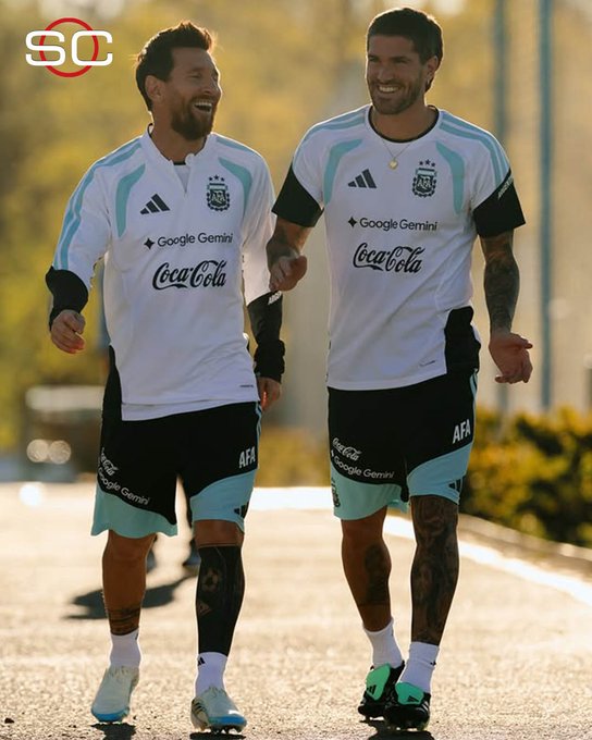 Lionel Messi and Rodrigo De Paul share a laugh during what appears to be an Argentina national team training session, wearing white kits featuring "Google Gemini" and "Coca-Cola" sponsorships alongside the AFA crest. The image captures the joyful camaraderie between the longtime midfield duo, visually embodying the post's sentiment that De Paul was the teammate who made Messi laugh the most during their time together with the "Scaloneta."