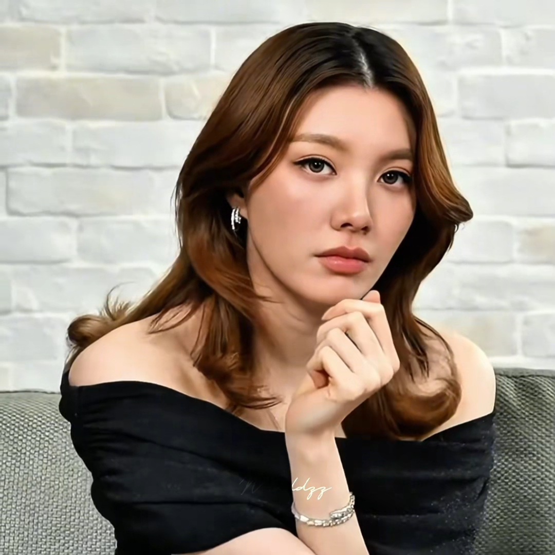 A woman with wavy brown hair wearing a black off-the-shoulder top poses thoughtfully with her hand near her chin in front of a white brick wall.