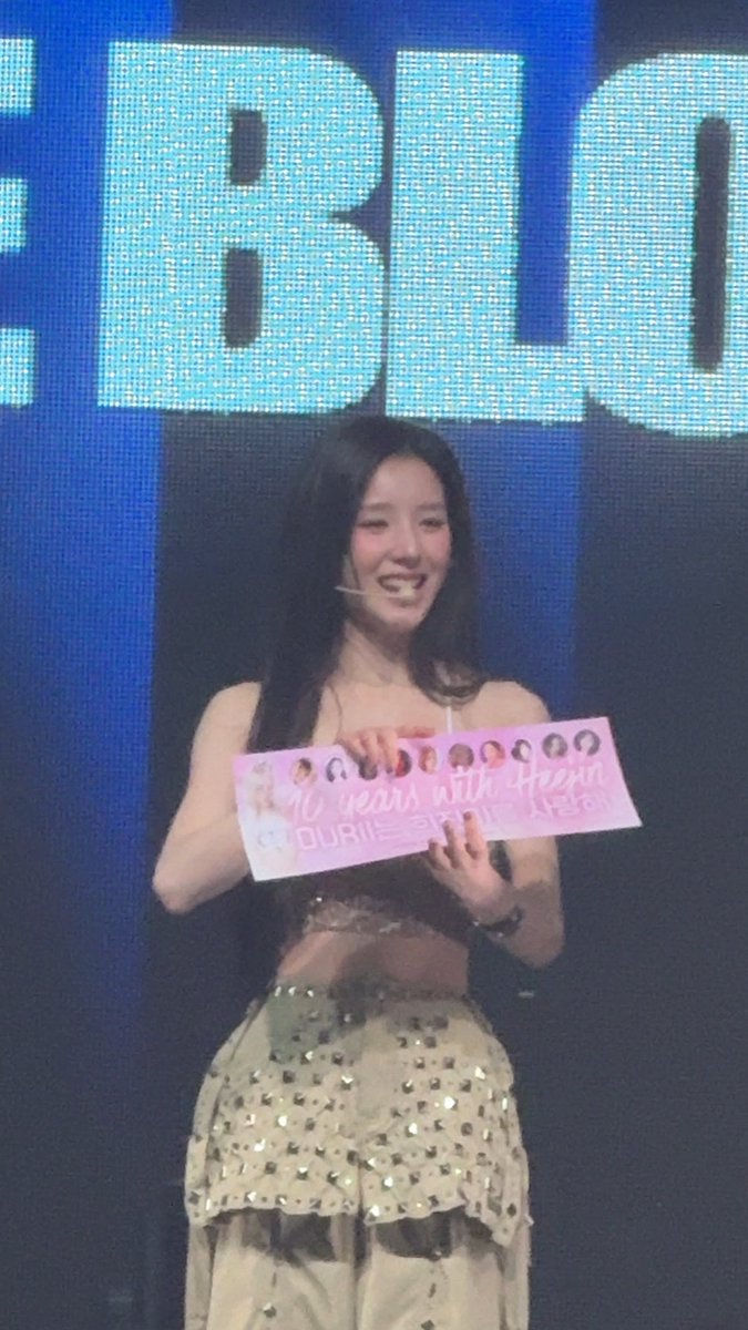 Heejin, a member of ARTMS, stands on stage at a Toronto concert beaming with a bright smile as she holds up a fan-made pink banner reading "10 years with Heejin" decorated with small portrait photos. The large blue screen behind her prominently displays "BLQ" (likely part of a larger text), with the hashtags #ARTMSinTORONTO and #ARTMS tying the moment to her milestone 10-year fan anniversary celebration.