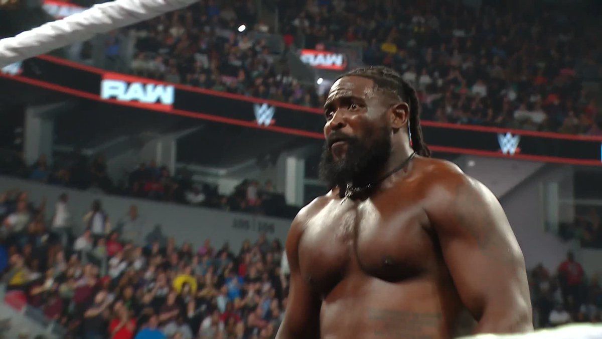 A WWE wrestler with dreadlocks and a beard stands shirtless in a wrestling ring, looking to the side, with a large arena crowd and a 'RAW' banner visible in the background.