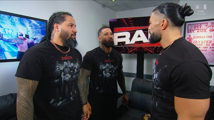 Three muscular men with dark hair and beards stand in a backstage area, all wearing matching black Bloodline t-shirts, with the man on the right facing the other two. Behind them, screens display the "RAW" WWE logo and a promotional poster mentioning a Saturday event featuring "SummerSlam" matches in Cleveland.