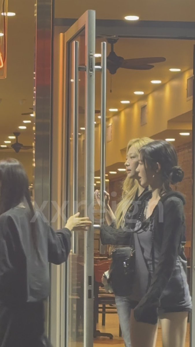 Two young women, identified by the post's hashtags as YUHA and STELLA of Hearts2Hearts, are seen entering a warmly lit establishment with glass doors, while a staff member in black holds the door open for them.