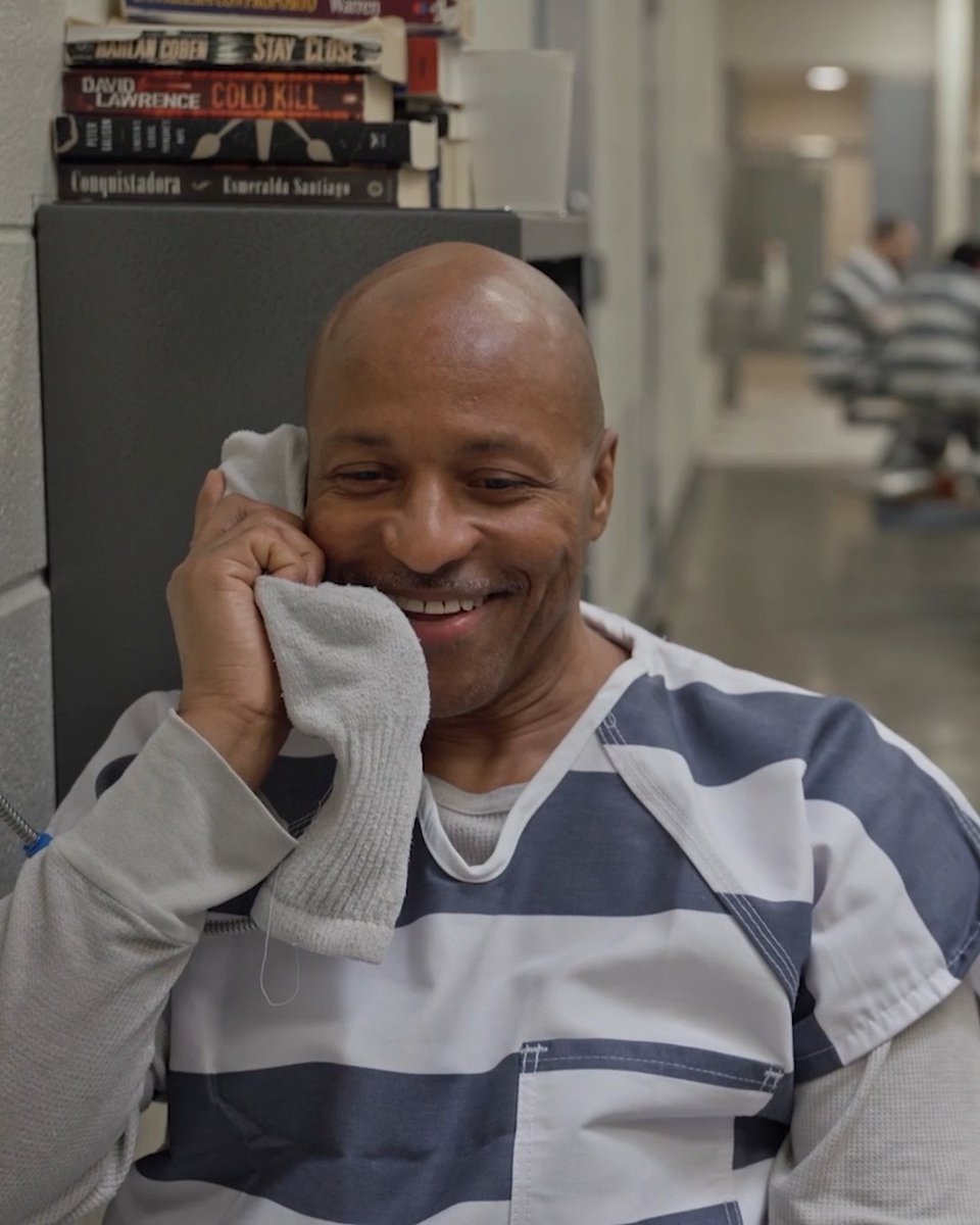 A smiling bald man in a striped prison uniform holds a white sock to his face during a phone call, with a stack of books behind him including titles like "Stay Close" by Harlan Coben, "Cold Kill" by David Lawrence, and "Conquistadora" by Esmeralda Santiago—visually setting the scene for his story as one of the few openly gay men in the facility. The contrast between his warm, genuine smile and the institutional surroundings captures a personal moment of self-acceptance within the #120HoursBehindBars experience.