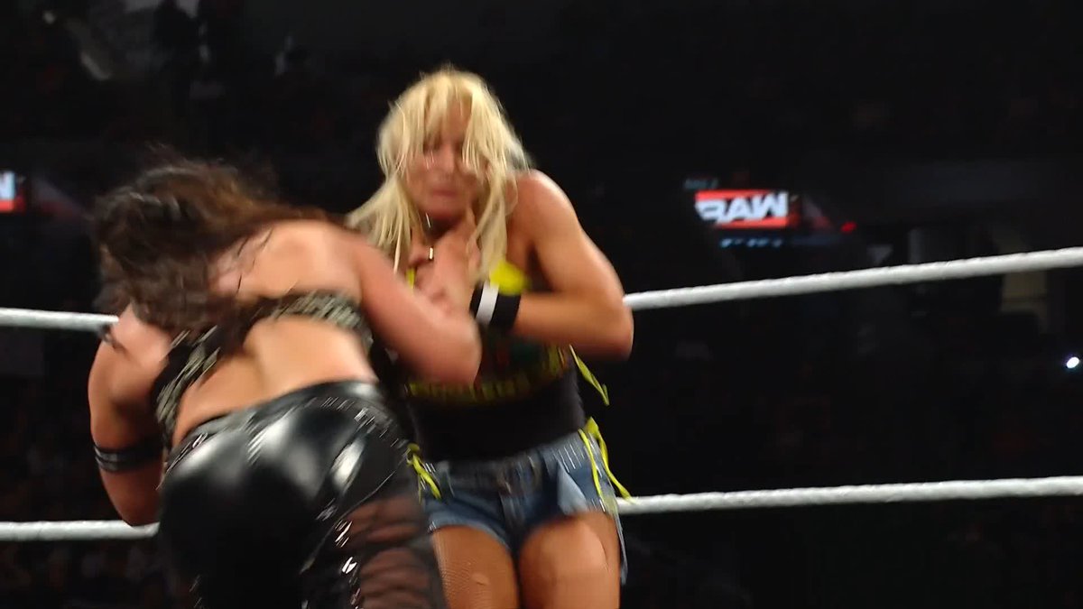 Two female wrestlers engage in close combat inside a WWE Raw ring, capturing a tense moment of physical confrontation. In the upper right, a screen shows the "RAW" logo, indicating the brand of the wrestling event.