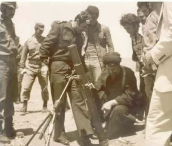 A sepia-toned historical photograph showing a group of soldiers in uniforms gathered around military equipment, with one seated soldier holding an object.