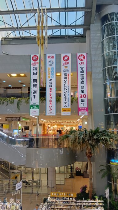 The interior of a Japanese shopping mall features hanging vertical banners with Japanese text, including '祝 30周年' (celebrating 30 years) for Fukuya department store, under a glass atrium ceiling.