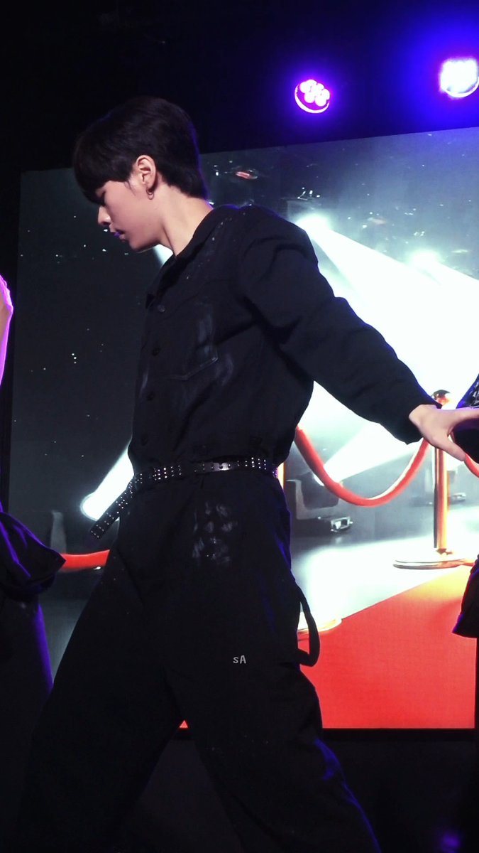 A young man in an all-black outfit with a studded belt poses on a stage with bright blue and purple lights, a red carpet, and a red velvet rope in the background. The watermark 'sA' is visible on his pants.
