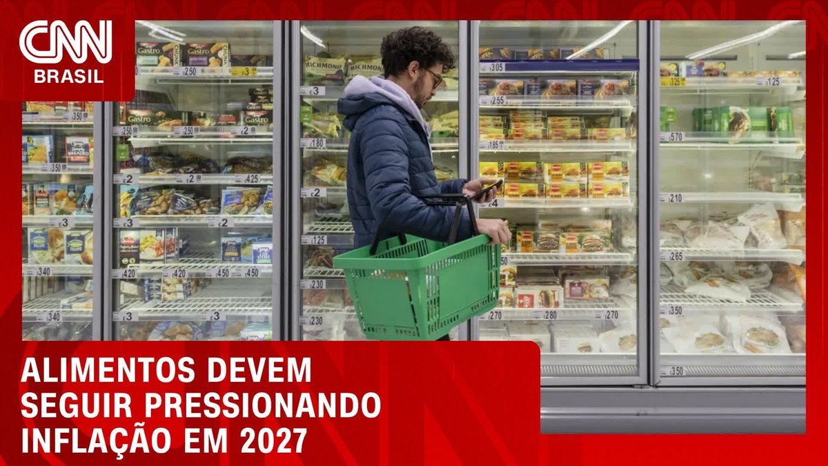 A man in a blue jacket holding a green shopping basket examines a product in front of a refrigerated grocery store section, with the CNN Brasil logo and headline "ALIMENTOS DEVEM SEGUIR PRESSIONANDO INFLAÇÃO EM 2027" (Food should continue to pressure inflation in 2027) displayed, illustrating the rising cost of groceries discussed in the post.