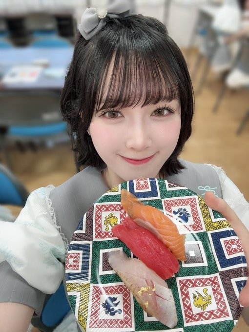 A young woman with short black hair and a small bow, wearing a ruffled gray and white outfit, smiling at the camera while holding a colorful patterned plate with three pieces of sushi.